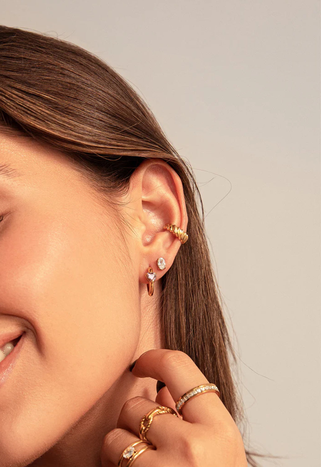Close-up of a person wearing gold earrings and rings on a neutral background