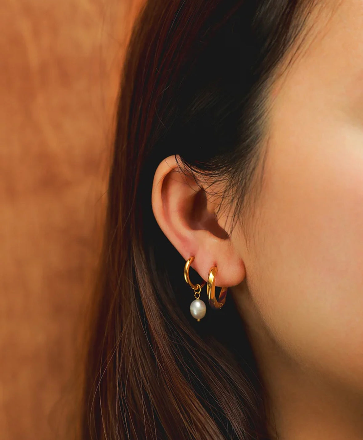 Close-up of an ear wearing gold hoop earrings with a pearl earring against a warm-toned background.