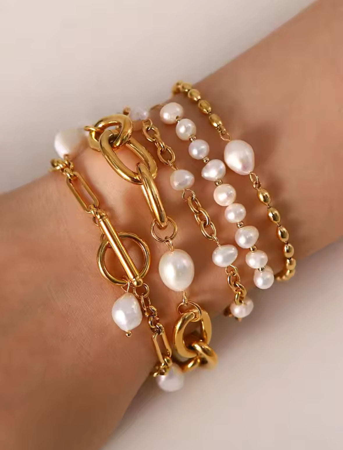 Gold chain and pearl bracelet on a wrist against a neutral background