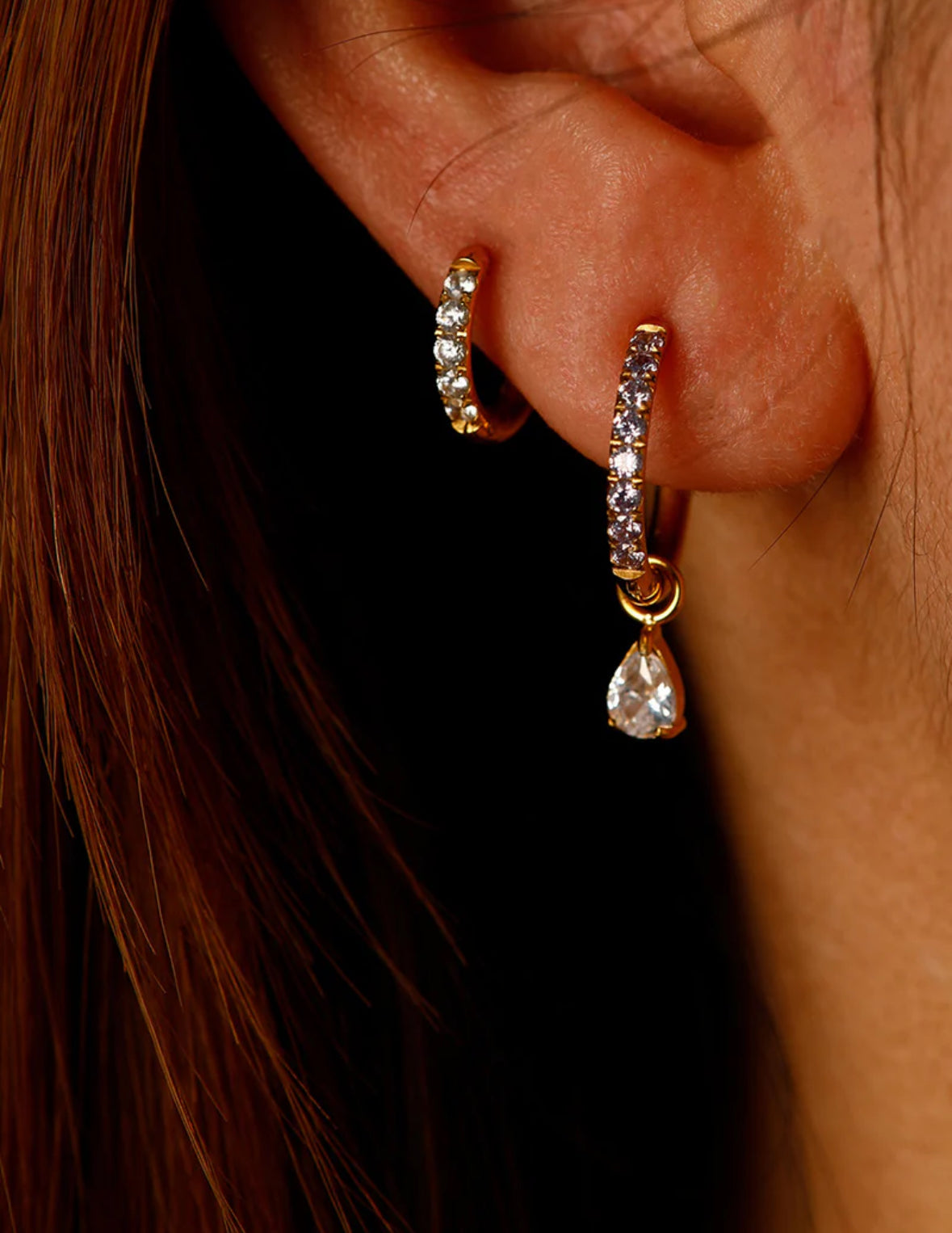 Close-up of an ear wearing gold hoop earrings with clear gemstones.
