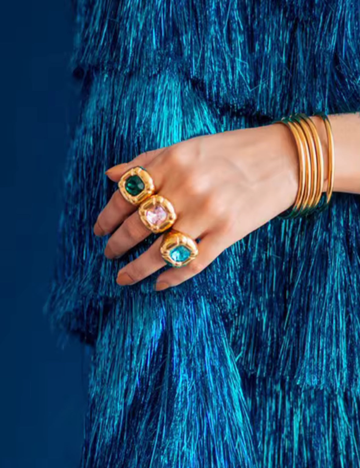 Hand wearing gold rings with gemstones against a blue textured background