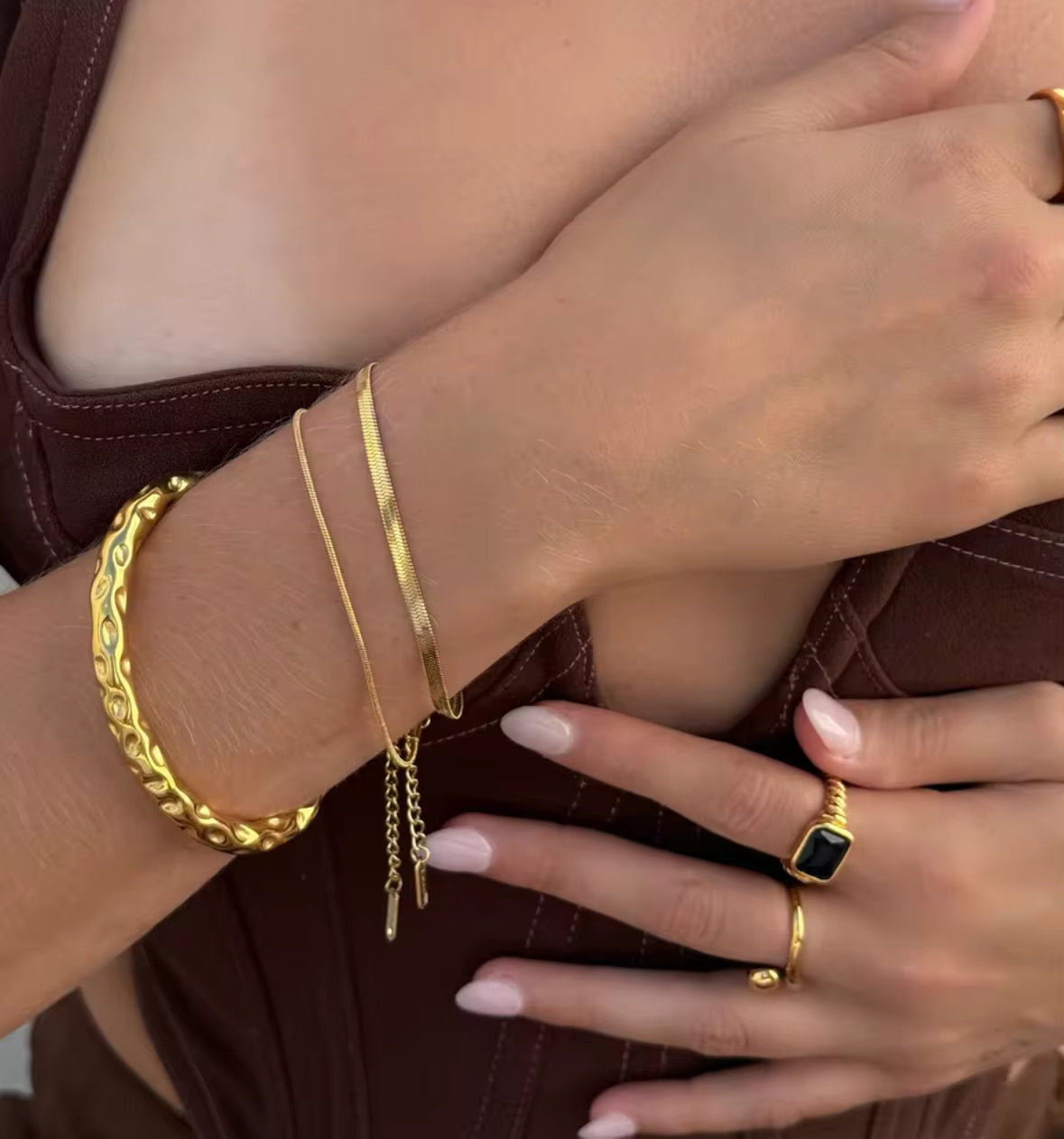 Gold bracelets and rings on a person's wrist and hand against a brown background