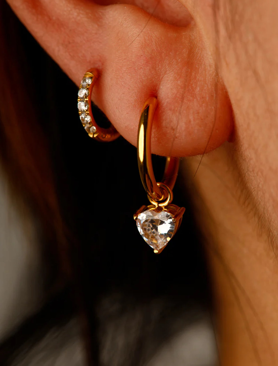 Gold hoop earrings with heart-shaped gemstones worn on an ear.