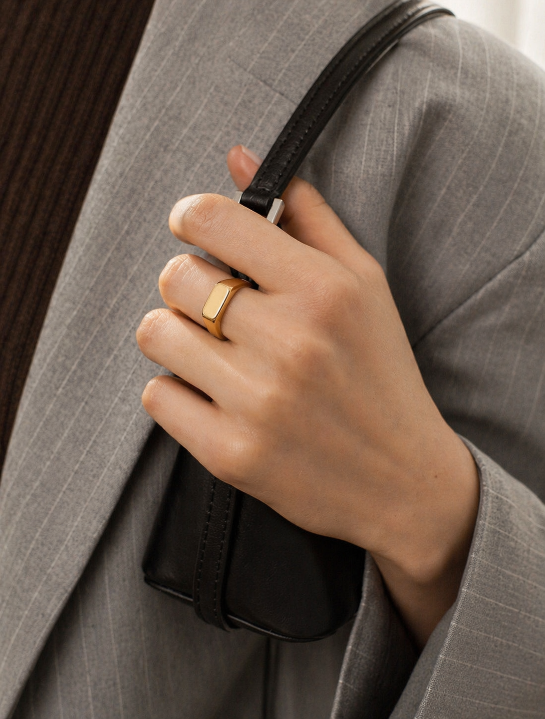 Hand holding a black handbag with a gold ring, wearing a gray suit.