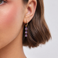 Close-up of a person wearing gold hoop earrings with purple gemstones against a neutral background