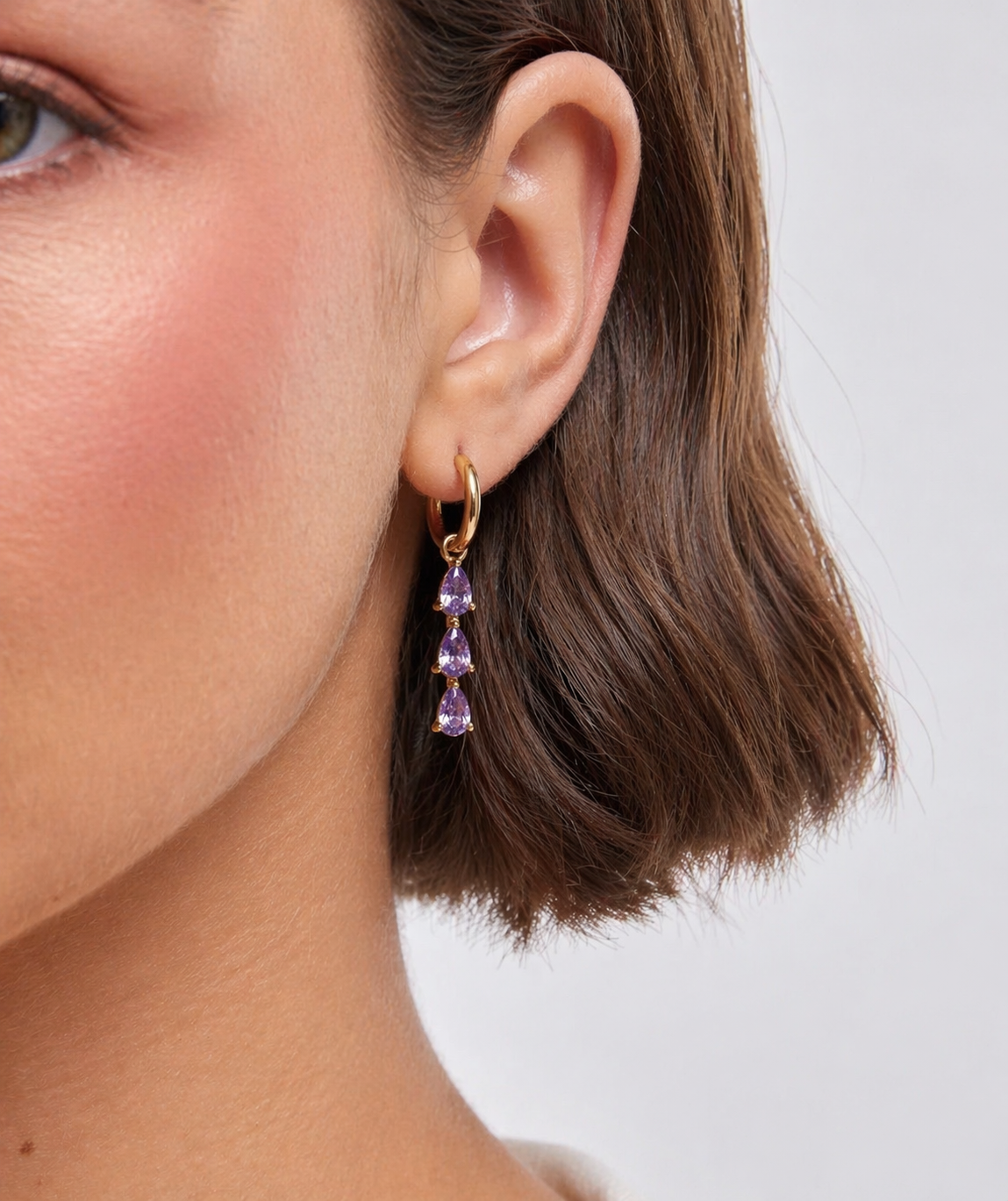 Close-up of a person wearing gold hoop earrings with purple gemstones against a neutral background