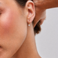 Close-up of a person wearing a gold earring with a clear gemstone on a neutral background