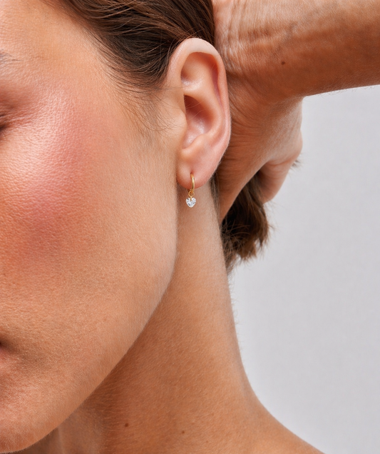 Close-up of a person wearing a gold earring with a clear gemstone on a neutral background