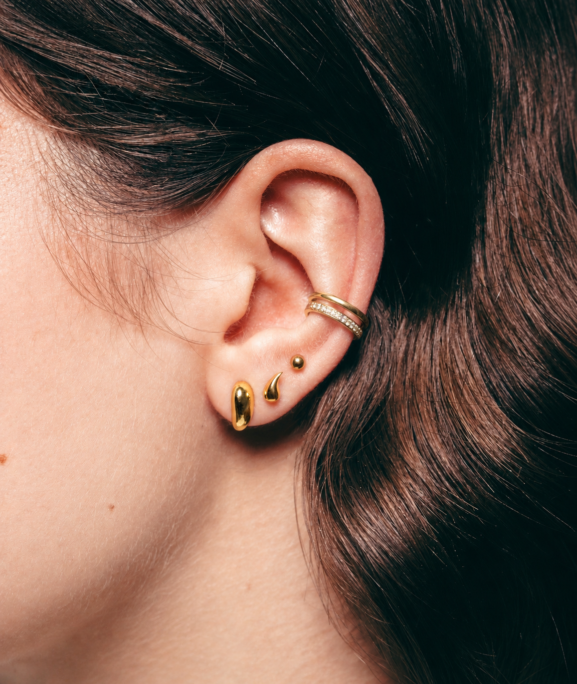 Close-up of an ear with gold earrings and a ring, with brown hair.