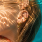Close-up of a person's ear with gold earrings underwater, with sunlight filtering through water.