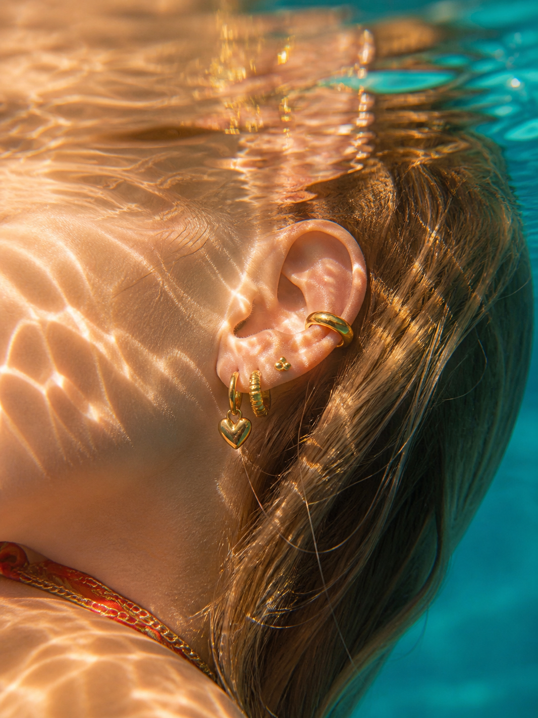 Close-up of a person's ear with gold earrings underwater, with sunlight filtering through water.