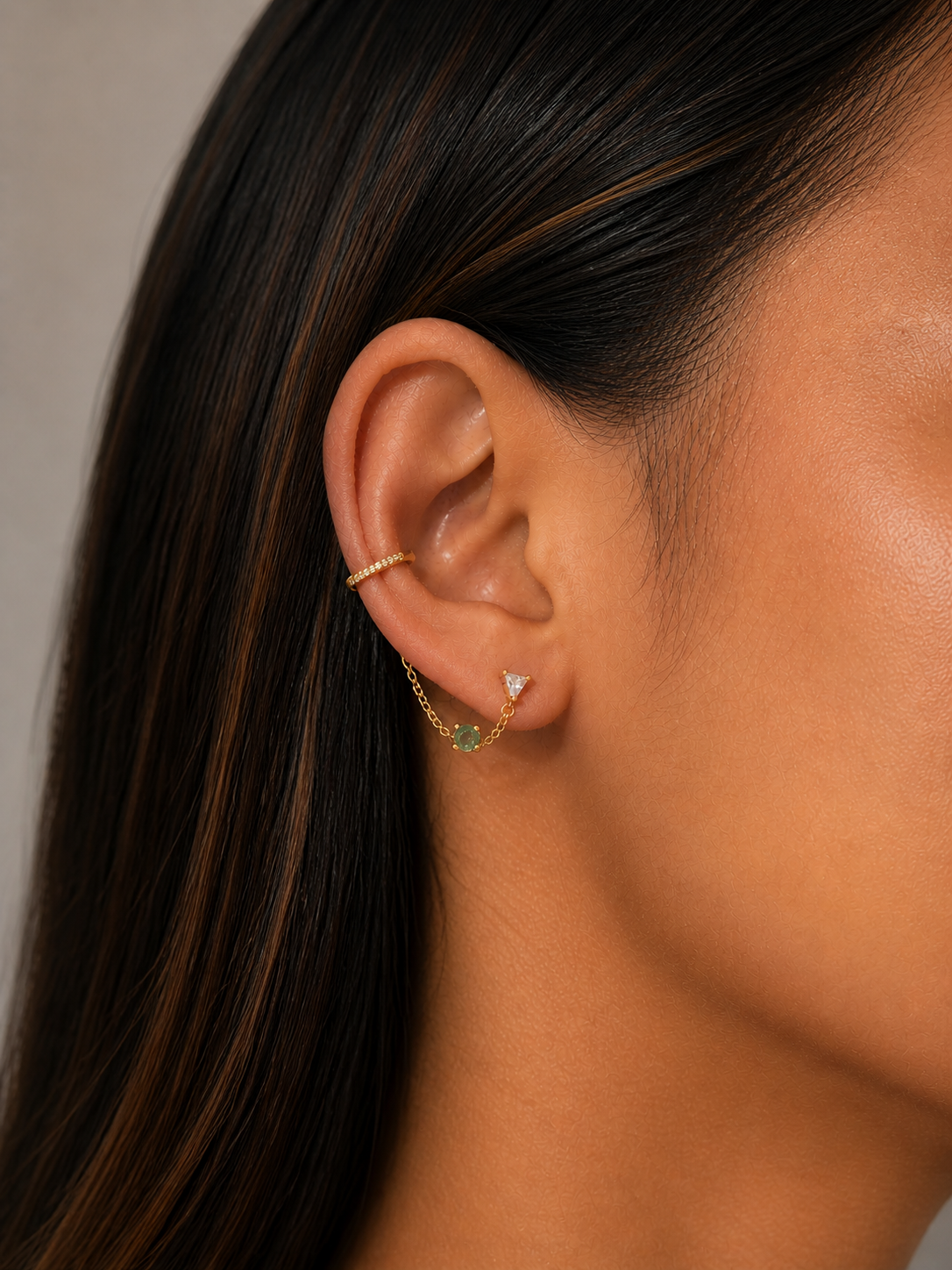 Close-up of an ear with a gold earring and green gemstone on a neutral background